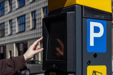 Şehrin caddesinde dokunmatik ekran parkmetresiyle etkileşime giren bir kadının eli. Modern terminalde mavi bir park yeri ve ödeme seçenekleri bulunmaktadır.. 