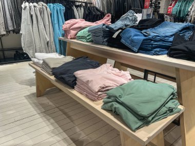 Modern bir perakende mağazasının içindeki ahşap masalara dizilmiş kot pantolonlu, düzgün katlanmış pastel ve nötr renkli tişörtler gündelik kıyafet organizasyonları ve mevsimsel trendleri gözler önüne seriyor.