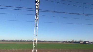 Demiryolu elektrik hatları düz bir tarla boyunca uzanır yeşil tarlalar ve açık mavi gökyüzünün altındaki uzak çiftlik evleri, taşımayı ve kırsal altyapıyı sembolize eder.