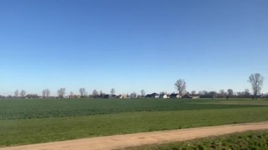 Tepedeki elektrik hatlarıyla ahşap elektrik direkleri toprak bir yolun yakınındaki çimenli tarlaları kaplıyor. Parlak, bulutsuz gökyüzü altında dağınık yapraksız ağaçlar ve kırsal evler var.