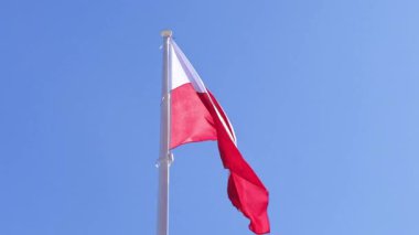 Uzun bir bayrak direğinde dalgalanan kırmızı ve beyaz Polonya bayrağı parlak ve berrak gökyüzüne karşı, güneşli bir bahar gününde vatanseverliği, ulusal gururu ve kimliği simgeliyor.