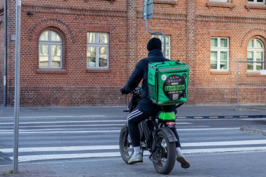 Poznan, Polonya - 8 Nisan 2025: Yeşil Uber 'li yemek kuryesi elektrikli bisiklet kullanıyor, tarihi tuğla bir binanın önündeki yaya geçidinde duruyor.