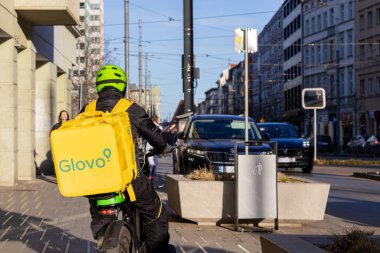 Poznan, Polonya - 8 Nisan 2025: Gıda kuryesi Glovo için yeşil kask takıyor ve büyük sarı bir sırt çantasıyla şehir caddesinde güpegündüz geziyor.