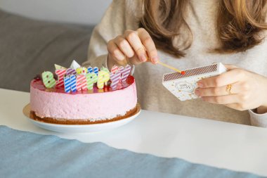 Bir kadının kırmızı jöleli pembe bir pasta üzerinde hafif çizgili doğum günü mumlarına kibrit çakarken, kapalı bir masada yumuşak ışıkla özel bir kutlama yaparken.