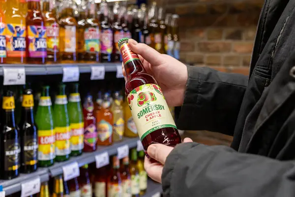 Poznan, Polonya - 8 Nisan 2025: Siyah ceketli bir adam renkli şişe içeceklerle dolu bir süpermarket rafının önünde bir şişe Somersby vişne ve elma şarabı tutuyor.