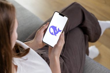 Poznan, Polonya 14 Nisan 2025: Kadın kullanıcı koltukta rahat bir şekilde oturuyor ve DeepSeek AI logosuyla akıllı telefon tutuyor, yapay zeka asistanlığını keşfediyor. 