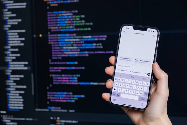 Poznan, Poland  April 14, 2025: Person holding smartphone with ChatGPT 4.5 chat open while coding on a computer, showcasing integration of AI tools into developer workflow 