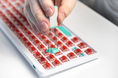 Kırmızı düğmeli mekanik bir klavyeden mavi bir tuşu kaldırmak için anahtar başlığı çekme aracı tutan bir elin yakın plan görüntüsü, DIY özelleştirme süreci ve teknik meraklısı kurulumu vurguluyor