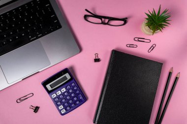 Pembe arka planda dizüstü bilgisayarı, kapalı siyah defteri, kalemleri, hesap makinesi, gözlüğü ve kırtasiyesi olan düzenli çalışma alanı, üretkenliği, planlamayı ve yaratıcılığı simgeliyor.