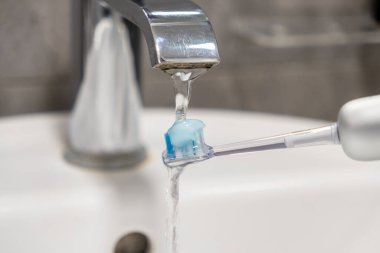 Elektrikli diş fırçasının kafasını lavabonun üzerindeki musluğun altından akan suyun altında diş macunuyla durulamak günlük ağız sağlığı için gerekli hazırlığı sembolize eder.