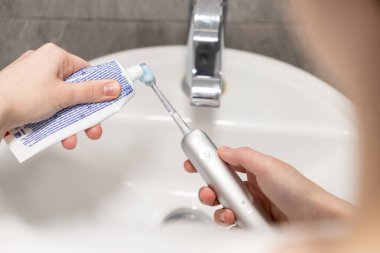 Bir insanın eli elektrikli diş fırçasının başına sabah rutinlerinin ve istikrarlı oral sağlığın önemini sembolize ederek lavabonun üzerine diş macunu sürer.