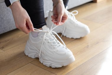 Eller, modern tıknaz spor ayakkabılarda beyaz ayakkabı bağcıklarını emniyete alırken ahşap zeminde rahat ve şık ayakkabılarla hazırlanma rutinini gösteriyor.