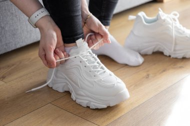 Kapalı mekanda beyaz bağcıklı spor ayakkabıları bağlayan ellerin detaylı görüntüsü, rahat spor ayakkabıları hazırlama rutinini gösteriyor.