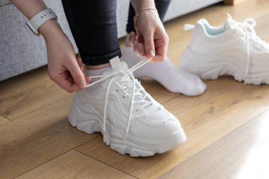 Modern spor ayakkabılarının bağcıklarını bağlayan eller ahşap zeminde, rahat ve aktif spor kıyafetlerle hazırlanmanın günlük rutinini gösteriyor.