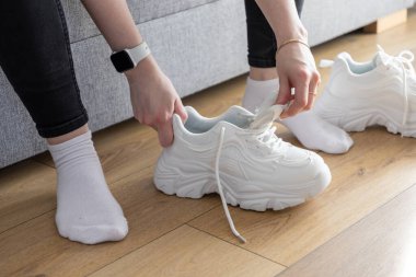 Modern spor ayakkabılarını kapalı alanda, ahşap zeminde çekerken ellerinizi kapayın, rahat spor ayakkabılarıyla hazırlanma rutinini gösterin.