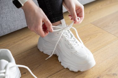 Kapalı mekanda beyaz bağcıklı spor ayakkabıları bağlayan ellerin detaylı görüntüsü, rahat spor ayakkabıları hazırlama rutinini gösteriyor.