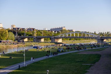 Poznan, Polonya - 25 Nisan 2025: Warta Nehri kıyısı parkında insanlar çimenli bir kıyıda dinleniyor ve açık bir gökyüzü altında bir köprü altında