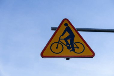 Kırmızı kenar ve siyah bisiklet sürücüsü simgesine sahip üçgen sarı uyarı işareti, yol kullanıcı güvenliğini destekleyen bisikletçiler hakkında trafik uyarısı niteliği taşıyor