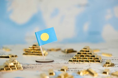 Küresel ekonomik eğilimleri, ulusal finans rezervlerini ve uluslararası ticaret politikalarını vurgulayan bulanık bir arka planda, küçük bir Palau bayrağı ve sayısız altın külçesinin yakın plan görüntüsü