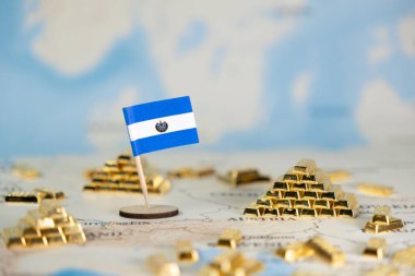 Bulanık bir dünya haritasında El Salvador 'un ulusal bayrağının ve minyatür altın külçelerinin sembolik yakın çekimi ulusal ekonomik büyümeyi ve küresel yatırımı geniş bir kopya alanı ile göstermektedir.