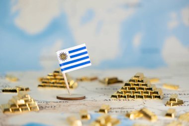 Küresel bir haritaya yerleştirilmiş minyatür bir Uruguay bayrağının ayrıntılı görüntüsü, çoklu altın külçeleri, ulusal ekonomik refahı sembolize eden, istikrarlı finansal kaynakları sembolize eden