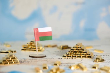 Umman bayrağının, minyatür altın külçeli piramitlerin yanında ülkenin finansal gücünü ve küresel ticaretini temsil eden antik bir dünya haritasına yakın plan görüntüsü.