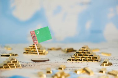 Küresel finans piyasaları, ekonomik potansiyeli ve uluslararası ticaret değerini tasvir eden, coğrafi haritaya saçılmış sayısız altın külçesiyle birlikte Türkmenistan bayrağının yakın görüntüsü