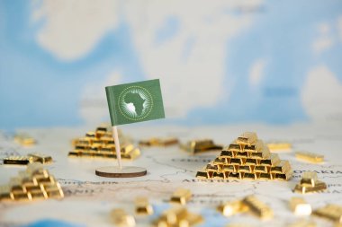 Minyatür Afrika Birliği bayrağı, kıtanın zengin kaynakları, ekonomik büyümesi ve küresel nüfuzunu gösteren sayısız altın külçelerle çevrili küresel bir haritaya belirgin bir şekilde yerleştirilmiştir.