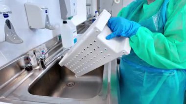 Mavi eldivenli ve yeşil koruyucu elbiseli bir tıp uzmanı paslanmaz çelik lavabodaki delinmiş bir tepsiyi titizlikle temizliyor ve güvenlik için gerekli sterilizasyonu vurguluyor.