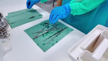 Parlak paslanmaz çelik cerrahi aletlerin yakından görüntüsü, makas ve kıskaçlar da dahil, nemli yeşil bir tıbbi paspasın üzerinde, steril uygulamalara ve tıbbi hijyene odaklandığını gösteriyor.