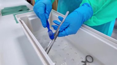 Tıp işçilerinin eldivenli elleri su dolu bir tepside cerrahi aletleri titizlikle temizler. Düzgün sterilizasyon ve hasta güvenliği için hayati önem taşır.