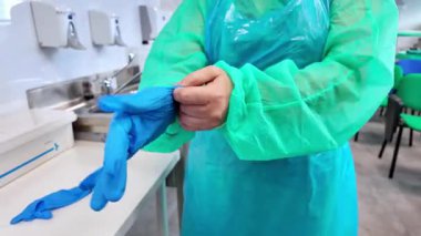 Yeşil koruyucu önlük ve mavi önlük giyerken mavi lateks eldiven takan ellerin yakın çekimi, tıbbi hijyen ve temel güvenlik önlemlerini temsil ediyor.