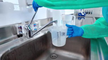 Eldivenli bir el, şeffaf bir ölçüm kabını temiz paslanmaz çelik bir lavaboda suyla doldurur ve bilimsel araştırmalar için gerekli olan sıvı kullanımını vurgular.
