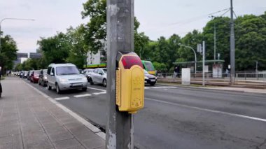 Bir kişinin eli, şehir altyapısı, sokak güvenliği ve etkin toplu taşıma etkileşimini gösteren bir kaldırımda parlak sarı bir yaya sinyal düğmesiyle temas kurar.
