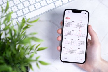 Poznan, Polonya - 10 Haziran 2025: Yeni iOS 26 ve Sıvı Cam tasarımı ile dolu bir 2025 takvimini parlak bir masanın üzerinde tutan akıllı telefon
