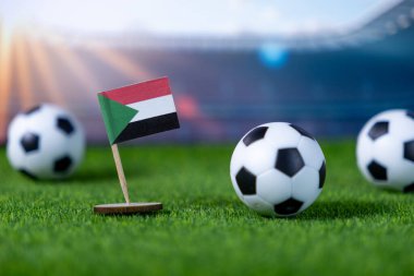 Küçük bir Sudan bayrağı ve minyatür futbol toplarının sentetik yeşil çimlerin üzerinde, yarışma sırasında bulanık bir stadyumun arka planına kurulmuş.