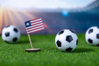 Canlı yeşil bir sahada minyatür futbol topları arasında dikilen Liberya bayrağının yakın plan görüntüsü ulusal spor hedeflerini ve küresel futbol yarışmasını temsil ediyor.