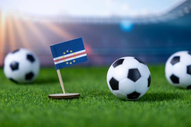 Cape Verde 'nin ulusal bayrağı ve futbol topları sentetik yeşil sahada, stadyumun arka planında, milli takım ruhunu ve küresel futbol etkinliklerini sergilemek için mükemmel.