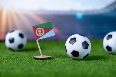 Close-up of a small Eritrean flag miniature and soccer balls on artificial grass, ready for an international football event in a blurred stadium background