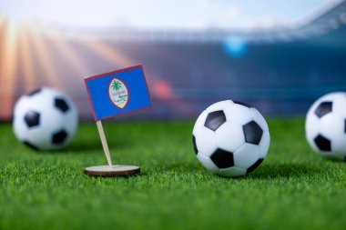 Ulusal amblemi ile küçük bir Guam bayrağının yakın çekimi, yapay çimlerin üzerinde üç futbol topuyla çevrili, küresel bir futbol maçına hazır.