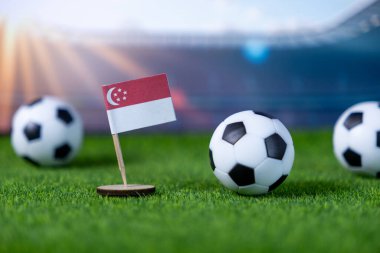 Stadyumun suni sahasında futbol topları bulunan küçük bir Singapur ulusal bayrağının yakın plan görüntüsü, spor etkinliği pazarlaması ve taraftar desteği için mükemmel.