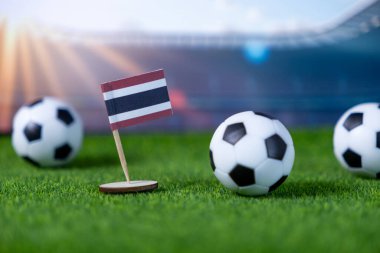 Minyatür Tayland bayrağı ve yeşil suni çimlerde klasik siyah beyaz futbol topları futbol tutkusunu, milli takım desteğini ve yaklaşan turnuvaları sembolize ediyor.