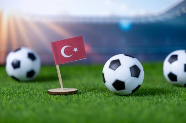 Canlı Türk bayrağı ve yapay yeşil çimenlikte klasik futbol topları, bulanık bir stadyumun arka planında, spor ve küresel futbolun heyecanını temsil eden ışıklarla çevrili.