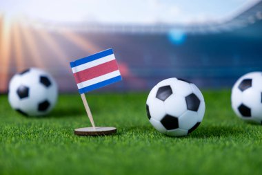 Minyatür futbol topları ve stadyum arka planında Kosta Rika bayrağı ulusal takım ruhunu, küresel futbol yarışmasını ve spor taraftarlarının heyecanını çağrıştırıyor.