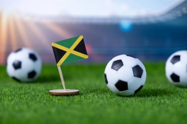 Yapay yeşil çimlerde küçük Jamaika bayrağı ve futbol topları. Bulanık stadyum ışıkları, uluslararası futbol etkinliklerini ve milli takım ruhunu simgeliyor.