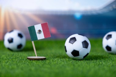 Meksika bayrağının ve sentetik yeşil çimlerin üzerindeki futbol toplarının yakın plan görüntüsü, bulanık bir stadyum zeminine karşı kurulmuş, küresel futbol turnuvalarını ve ulusal gururu vurguluyor.