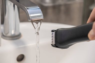 Çağdaş bir banyo musluğunun altında elektrikli tıraş makinesinin kafasını durulayan bir elin yakın plan görüntüsü.