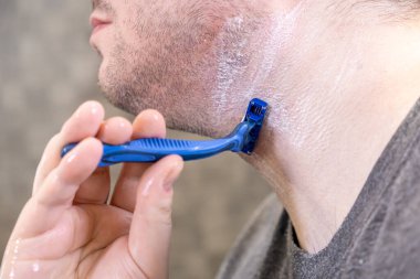 Mavi tek kullanımlık tıraş bıçağı tutan bir adamın elinin yakın çekimi, sakalları etkili bir şekilde temizler ve kişisel hijyen rutini sırasında temiz bir görünüm sağlar.