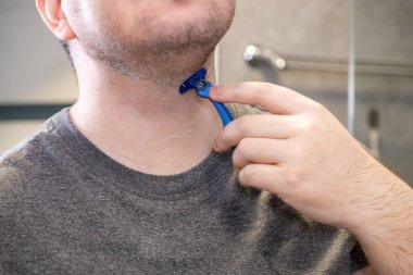 Bir adamın elinin tek kullanımlık mavi bir jileti tutarken yakın plan görüntüsü, ferahlatıcı bir kişisel bakım sırasında ustalıkla boynundaki sakalları çıkarıyor.