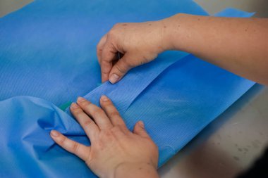 İnsan ellerinin dikkatlice katlanması, dokumasız steril ambalaj malzemesi, kontrollü bir tıbbi ortamda cerrahi aletlerin hijyenik ambalajının sağlanması.